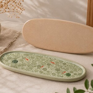 Peut inclure: Un plateau ovale en céramique avec un vernis vert clair orné de cercles superposés, de petits poissons rouges et de motifs circulaires vert foncé. Derrière se trouve une pièce ovale en céramique beige unie de même taille et forme.