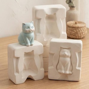 Könnte beinhalten: Eine hellblaue Keramikkatzenfigur sitzt auf einer rechteckigen, cremeweißen Gipsform. Zwei weitere rechteckige Gipsformen sind vorhanden, eine mit einer vertieften Katzensilhouette und die andere mit einem erhabenen Katzenrelief auf der Vorderseite.