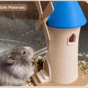 May include: A grey hamster drinking from a clear tube attached to a beige windmill-shaped water dispenser with a blue roof, a small door, steps, and a tiny potted plant. The text "Safe Materials" is visible in the upper left corner of the image.