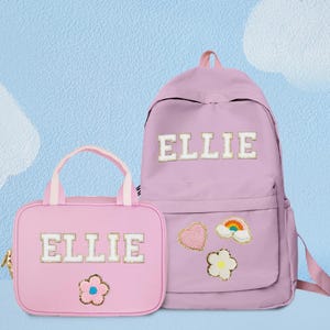 Könnte beinhalten: Ein passendes Set aus rosa Taschen, einem Rucksack und einer Tragetasche. Beide Taschen tragen den Namen "ELLIE" in weißen Blockbuchstaben. Der Rucksack hat eine Vordertasche und dekorative Aufnäher mit Herz, Regenbogen und Blume.