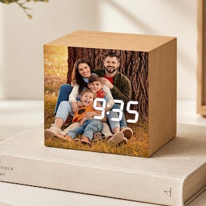Könnte beinhalten: Ein würfelförmiger digitaler Wecker mit heller Holzmaserung. Die Vorderseite zeigt ein Farbfoto einer vierköpfigen Familie, die im Freien vor einem Baumstamm sitzt, mit hellweißen digitalen Ziffern, die die Uhrzeit 9:35 anzeigen.