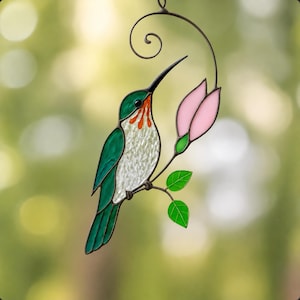 May include: A stained glass suncatcher featuring a hummingbird with green wings, a white textured chest, and orange throat feathers. The bird is perched on a stem with two green leaves and a single pink flower bud, framed by a dark metal scroll.