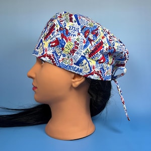 May include: A white scrub cap with a colorful comic book design featuring red, blue, and yellow accents. The cap is adorned with phrases like "Hero", "All American", and "Man of Steel". The cap is tied at the back.