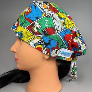 May include: A colorful scrub cap featuring comic book panels with Superman, Wonder Woman, and other DC Comics superheroes. The cap is made of fabric and has a tag that reads "DOUBLE".
