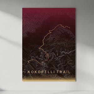 May include: A framed topographic map print of the Kokopelli Trail and Porcupine Rim. The map features a gradient of dark red to gold, with the trail highlighted in gold. The print is displayed on a white wall with a shadow.