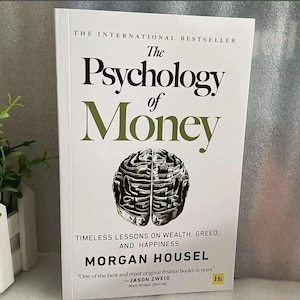 Puede incluir: Un libro blanco con el título "The Psychology of Money" en texto negro y verde. La portada del libro presenta una ilustración con forma de cerebro hecha de billetes de dólar. El libro es de Morgan Housel y se describe como "Timeless Lessons on Wealth, Greed, and Happiness". Una cita de Jason Zweig del Wall Street Journal está incluida en la portada.