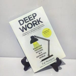 Könnte beinhalten: Ein Buch mit dem Titel "Deep Work" von Cal Newport wird auf einem schwarzen Ständer präsentiert. Das Buchcover ist weiß mit schwarzem Text und einer Grafik einer Schreibtischlampe. Der Untertitel lautet "Regeln für fokussierten Erfolg in einer abgelenkten Welt".