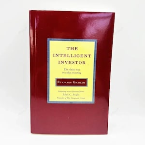 Op de afbeelding: Een hardcover boek getiteld "The Intelligent Investor" van Benjamin Graham. Het boek heeft een donkerrode kaft met een geel en blauw omkaderde titel. De tekst op de kaft bevat "The classic text on value investing" en de naam van de auteur van het voorwoord, John C. Bogle.