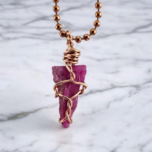 May include: A handmade necklace with a raw, deep pink ruby gemstone pendant. The ruby is wrapped in copper wire and hangs from a copper ball chain. The pendant has a unique, natural shape, highlighting the stone's texture and colour.