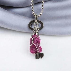 May include: A silver key necklace with a pink gemstone wrapped in wire. The key is attached to a silver chain.