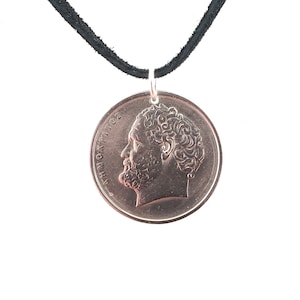 May include: A silver coin pendant with a profile of a man with curly hair, the coin has the text "ΑΥΤΟΚΡΑΤΟΡ" around the edge, hanging from a black cord.