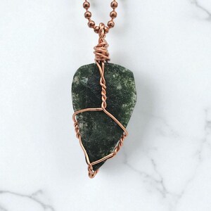 May include: A handcrafted pendant necklace featuring a dark green stone wrapped in copper wire. The stone has a natural, textured appearance and is suspended from a copper ball chain. The pendant is a unique piece of jewelry.