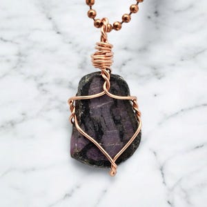 May include: A handcrafted necklace featuring a dark purple stone pendant wrapped in copper wire. The stone has black veining and is suspended from a copper ball chain. The wirework is intricately designed, showcasing the natural beauty of the stone.