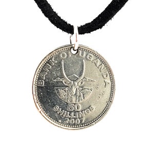 May include: A silver coin necklace with a black suede cord. The coin features an image of an antelope with large horns and the text "BANK OF UGANDA 50 SHILLINGS 2007".