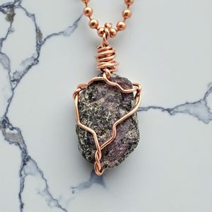May include: A copper wire-wrapped pendant necklace featuring a rough, dark purple stone. The stone is suspended from a copper ball chain. The wire is intricately wrapped around the stone, creating a unique, handcrafted jewelry piece.