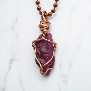 May include: A handcrafted necklace featuring a raw, deep magenta ruby gemstone wrapped in copper wire. The pendant hangs from a copper ball chain. The ruby is a rough, natural stone, showcasing its unique texture and color variations. The wirework adds an artistic touch.