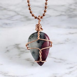 May include: A teardrop-shaped pendant necklace featuring a polished stone with gray, white, and burgundy hues. The stone is wrapped in copper wire and suspended from a copper ball chain. The necklace is displayed against a marble background.