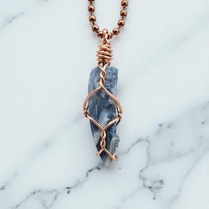 May include: A handcrafted pendant necklace featuring a blue and gray stone wrapped in copper wire. The stone is suspended from a copper ball chain. The pendant is displayed against a white marble background.