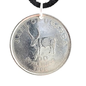 May include: A silver coin pendant with a black cord. The coin features an image of an antelope with large horns and the text "BANK OF UGANDA 100 SHILLINGS 1998".