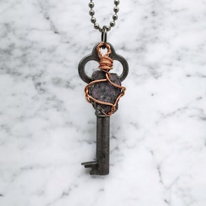 以下が含まれることがあります： ヴィンテージスタイルの鍵ペンダント。濃いグレーの仕上げで、紫色の原石が銅線で包まれ、鍵の頭に取り付けられています。ペンダントはシルバーカラーのボールチェーンから吊り下げられており、ユニークな手作りのネックレスになっています。