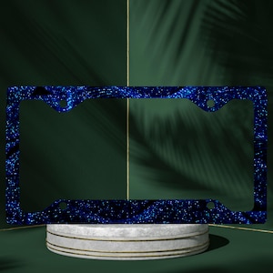 May include: A blue license plate frame with a dark blue and white glitter design. The frame has a rectangular shape with rounded corners and pre-drilled holes for mounting. The background is a dark green with a white and gold pedestal.