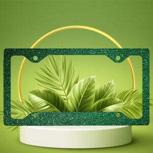 May include: A green glitter license plate frame with a gold ring and green leaves on a white pedestal. The frame has four holes for mounting. The background is a gradient of green.
