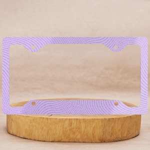 May include: A license plate frame with a wavy pattern in light purple. The frame has a repeating dot design and is displayed on a wooden surface.