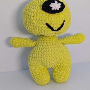 May include: A crocheted yellow plush toy with a black eye patch and a white flower on the patch. The toy has a smiling mouth and outstretched arms.