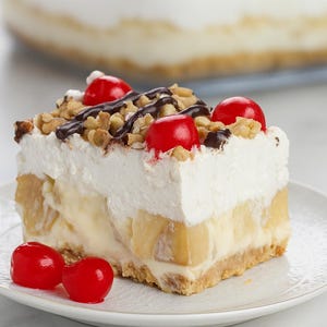 Puede incluir: Una porción de postre de crema de plátano sobre un plato blanco, cubierta con crema batida, salsa de chocolate, nueces picadas y cerezas rojas. El postre tiene capas de plátanos, crema y una base de galleta. Otro postre está en el fondo.