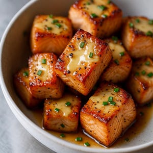 Può includere: Una ciotola bianca piena di bocconcini di salmone cotti. Il salmone è marrone dorato con una glassa lucida e guarnito con erba cipollina verde tritata. Il piatto è presentato su una superficie grigio chiaro.