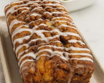 May include: A loaf of cinnamon swirl bread with white icing drizzled on top, presented on a rectangular plate. A bowl of green apples sits in the background. The bread has a golden-brown color.