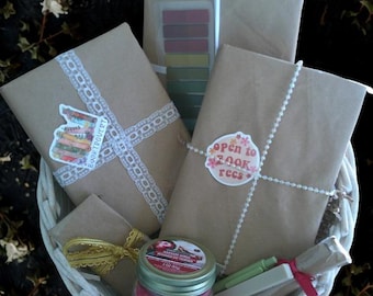 May include: A white wicker basket containing wrapped books, a pink candle, and stationery. The books are wrapped in brown paper and decorated with lace, stickers, and string. The candle jar has a silver lid and a label. The text "open to book recs" is visible.