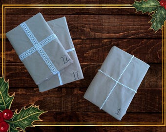 May include: Three wrapped gifts on a wooden surface, possibly for an advent calendar. The gifts are wrapped in gray paper and tied with white ribbon. Numbers 1, 17, and 22 are written on the packages. Holly and berries decorate the corners.