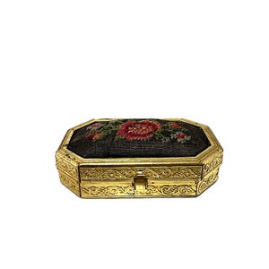 Könnte beinhalten: Eine antike, achteckige Schmuckschatulle mit einem goldfarbenen Metallrahmen. Der Deckel ist mit schwarzem Stoff bestickt, der rote und rosa Blumen sowie grüne Blätter zeigt. Die Schatulle hat verzierte, gravierte Details.