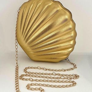 Może przedstawiać: Złota torebka w kształcie muszli z łańcuszkiem. Torebka ma teksturowaną powierzchnię z promienistymi liniami, a łańcuszek jest również złoty. Unikalny dodatek.