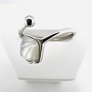 Könnte beinhalten: Ein silberfarbener Ring mit einem muschelförmigen Design und einem kleinen Kugelakzent. Der Ring hat eine perlmuttweiße, fächerförmige Einlage und ein gebogenes Band. Der Ring wird auf einer weißen Oberfläche präsentiert.