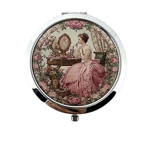Könnte beinhalten: Ein runder, silberner Kompaktspiegel mit einer Vintage-Illustration einer Frau, die sich schminkt. Das Bild zeigt eine Frau in einem rosa Kleid an einem Schminktisch, umgeben von rosa Rosen. Der Spiegel hat eine Klappöffnung und einen Druckknopfverschluss.