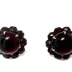 Könnte beinhalten: Ein Paar Ohrstecker mit einem großen, dunkelroten, im Cabochon-Schliff geschliffenen Edelstein in der Mitte, umgeben von einem gewellten Rand aus kleineren, facettierten dunkelroten Steinen in einer Metallfassung mit sichtbaren Krappen.