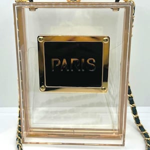 Könnte beinhalten: Eine transparente Acryl-Handtasche in Form einer Parfümflasche mit einer goldenen Kettenriemen und einer schwarzen Plakette mit dem Wort "PARIS" in goldenen Buchstaben.