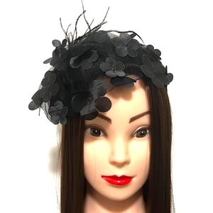Può includere: Copricapo nero con base in rete, decorato con fiori di tessuto e piume sottili. I fiori sono disposti sulla parte superiore, creando un effetto strutturato. Questo accessorio è adatto per occasioni speciali.