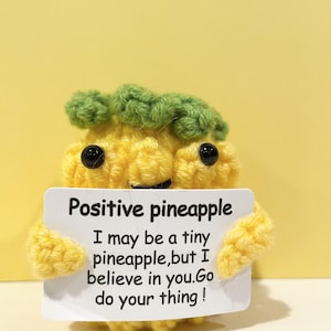 May include: A crocheted yellow pineapple figure with green leaves and black eyes. The pineapple holds a white sign that reads "Positive pineapple. I may be a tiny pineapple, but I believe in you. Go do your thing!"