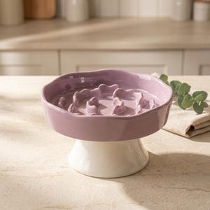 May include: A ceramic slow feeder bowl featuring a lavender-colored basin with an internal maze-like structure, supported by a solid white pedestal base. The bowl has a smooth, glossy finish and a wide, circular rim for holding food or water.