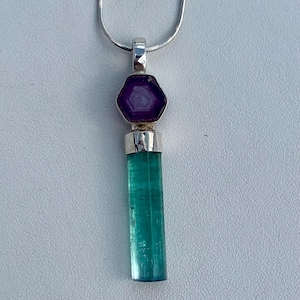 May include: A silver necklace featuring a pendant with a hexagonal purple stone set above a long, rectangular, translucent green tourmaline crystal. The pendant is suspended from a thin silver snake chain, showcasing a clean, metallic setting.