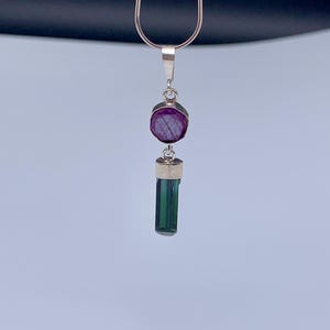 May include: A silver necklace featuring a pendant with a round, faceted deep pink gemstone positioned above a vertical, rectangular, dark green crystal. The pendant is suspended from a thin, smooth silver chain against a plain background.