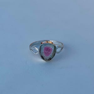 Puede incluir: Anillo de plata con una piedra preciosa de turmalina sandía. La piedra preciosa tiene un centro rosa, una banda blanca y un borde exterior verde. La banda del anillo tiene un diseño sencillo con recortes decorativos.