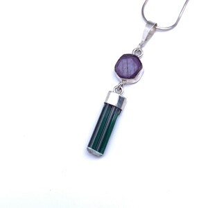 May include: A silver pendant necklace featuring a faceted, hexagonal deep purple stone set in a silver bezel, connected to a long, cylindrical, dark green tourmaline crystal capped in silver, suspended from a thin, flexible silver snake chain.
