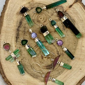 May include: A selection of gemstone pendants and earrings with silver settings, presented on a wooden surface. The gemstones are in various colours, including green, purple, and pink. The earrings have a hook design. The pendants and earrings are of different shapes and sizes.