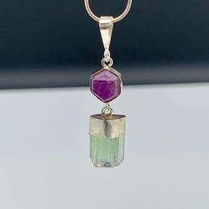 May include: A pendant necklace featuring a hexagonal deep pink ruby gemstone set in silver, suspended above a vertical, raw, light green tourmaline crystal encased in a silver cap. The pendant hangs from a thin, metallic silver snake chain.