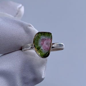 Puede incluir: Un anillo de plata con una piedra preciosa de turmalina sandía en bruto y asimétrica. La piedra muestra un degradado de color distintivo, que transita desde un borde exterior verde vibrante hacia un centro rosa suave. El aro tiene un acabado pulido.