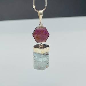 Può includere: Una collana in argento con un ciondolo composto da una pietra preziosa esagonale rosa scuro sospesa sopra un cristallo rettangolare azzurro. Il ciondolo è fissato a una sottile catena d'argento, mostrando un design a strati con due pietre.